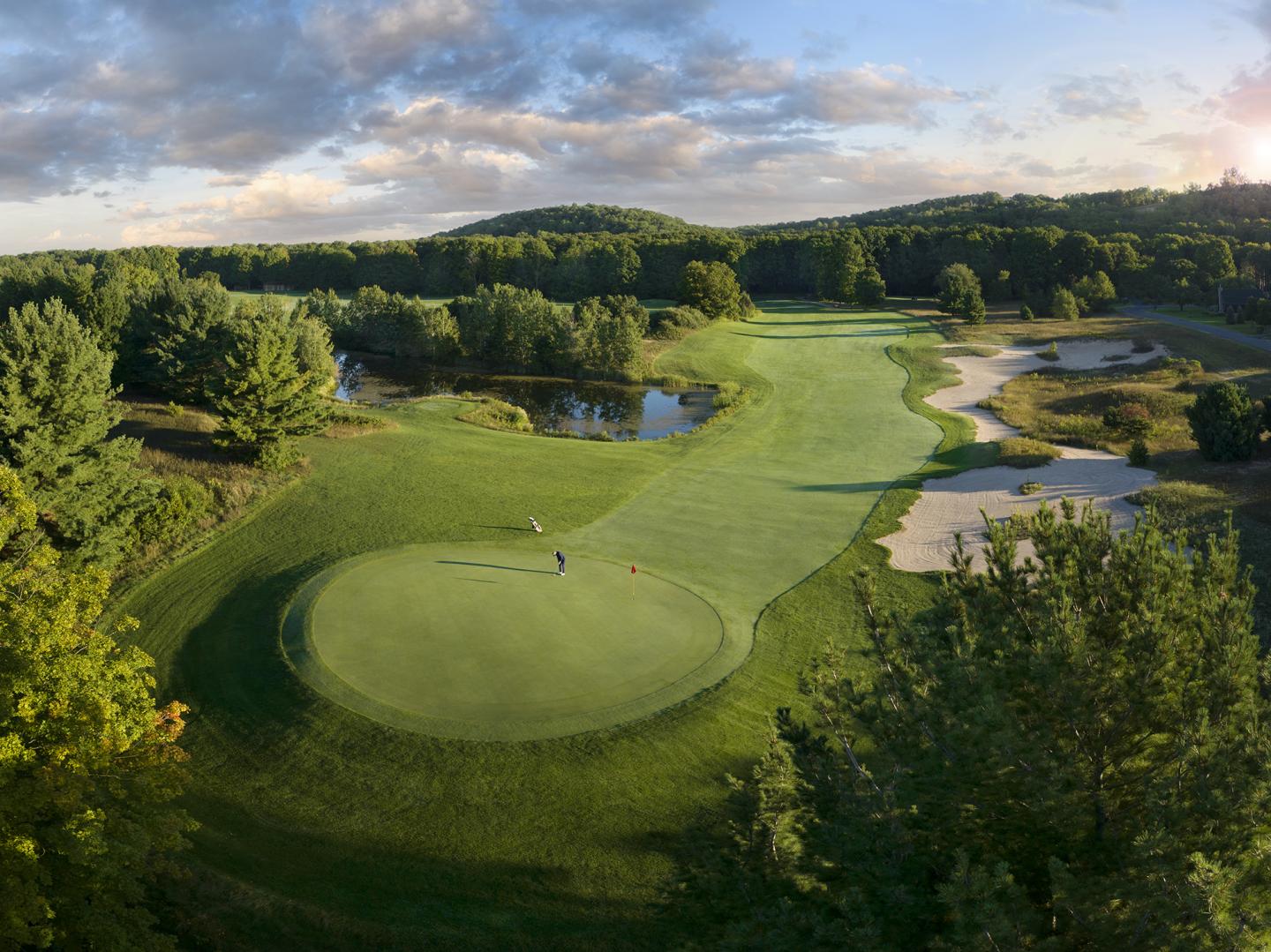 Golf course with lush green fairways, sand traps, and surrounding trees under a cloudy sky.