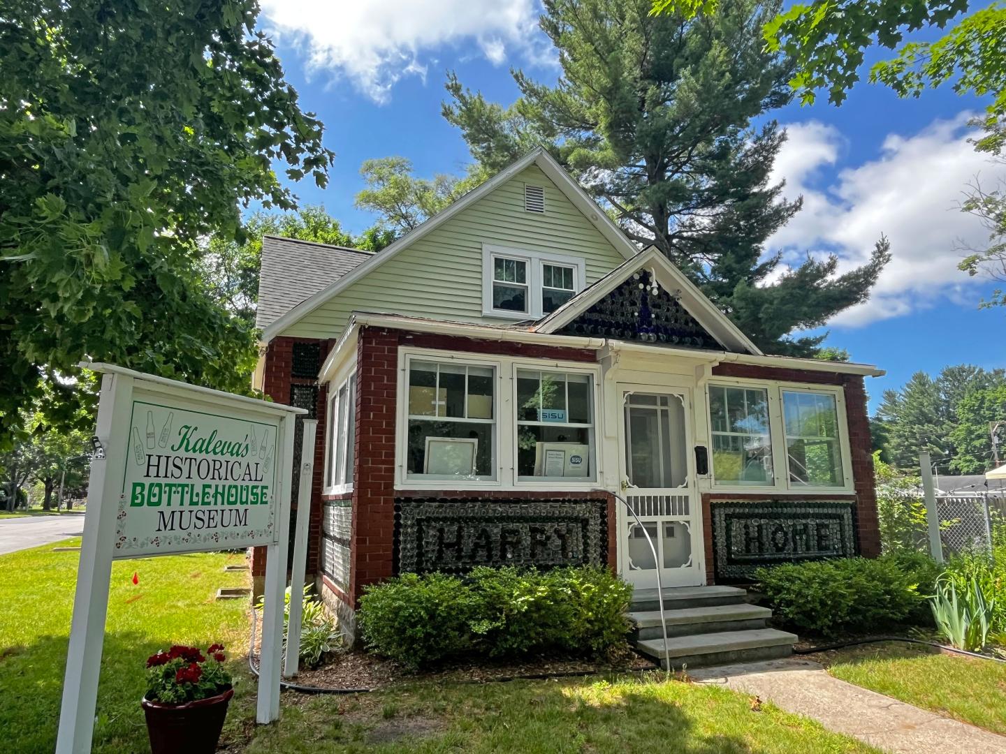 White house with a porch and green sign, surrounded by trees and grass.