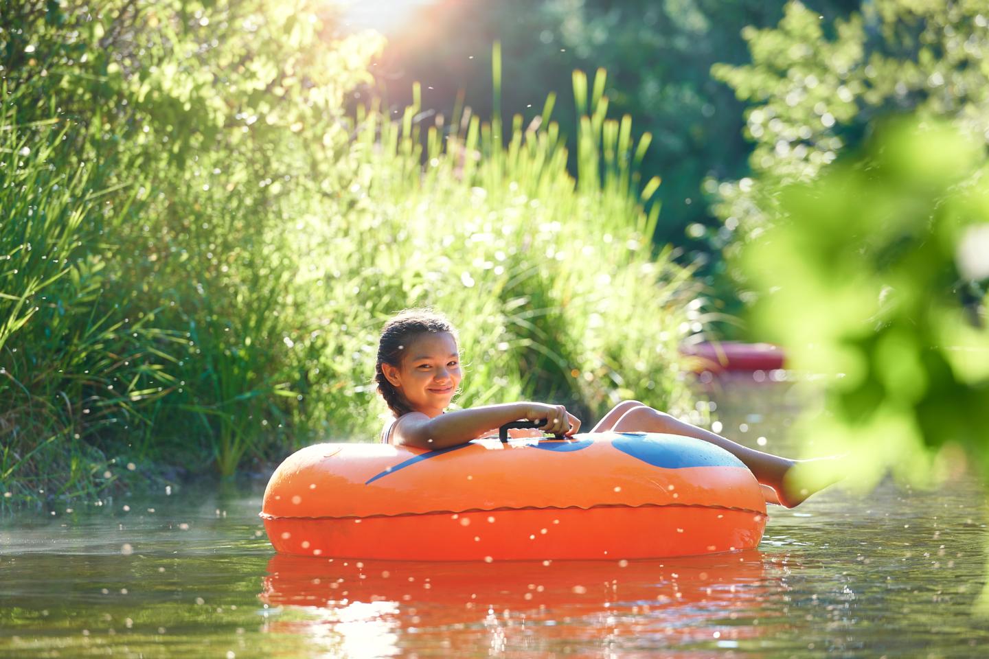 Child smiling on an orange float in a sunlit lake with lush greenery.