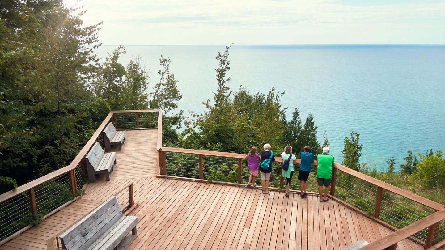 Four people stand on a wooden deck overlooking a blue lake and trees.