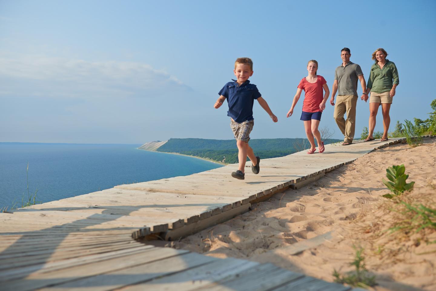 Family walking on a boardwalk by the sea under a clear blue sky.