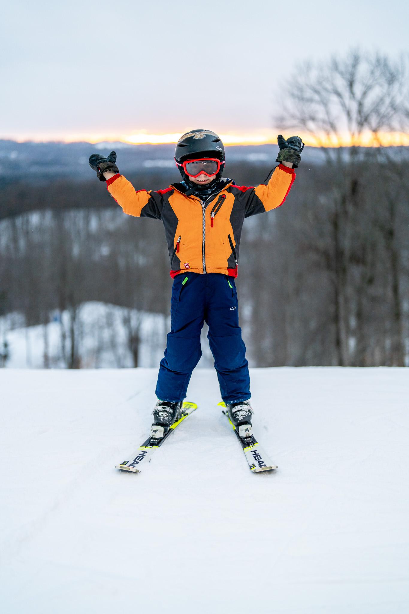 Child skiing in winter gear, arms raised, snowy landscape and sunset background.