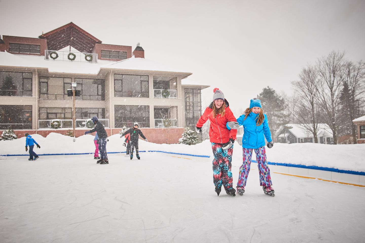 People ice skating near a snowy building, wearing colorful winter clothing.