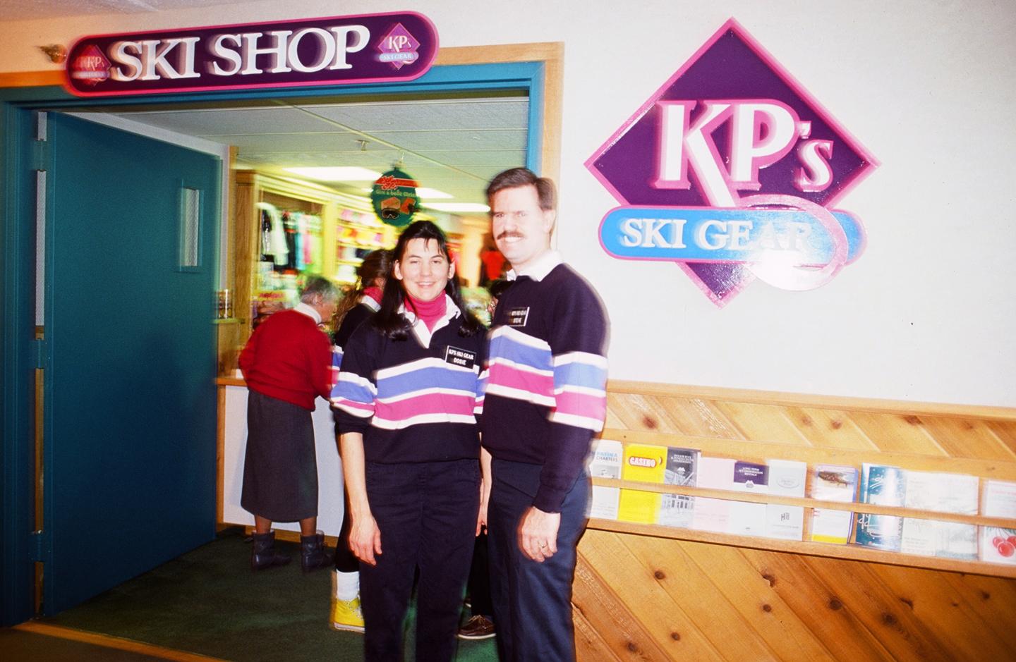Two people smiling in front of a ski shop entrance.