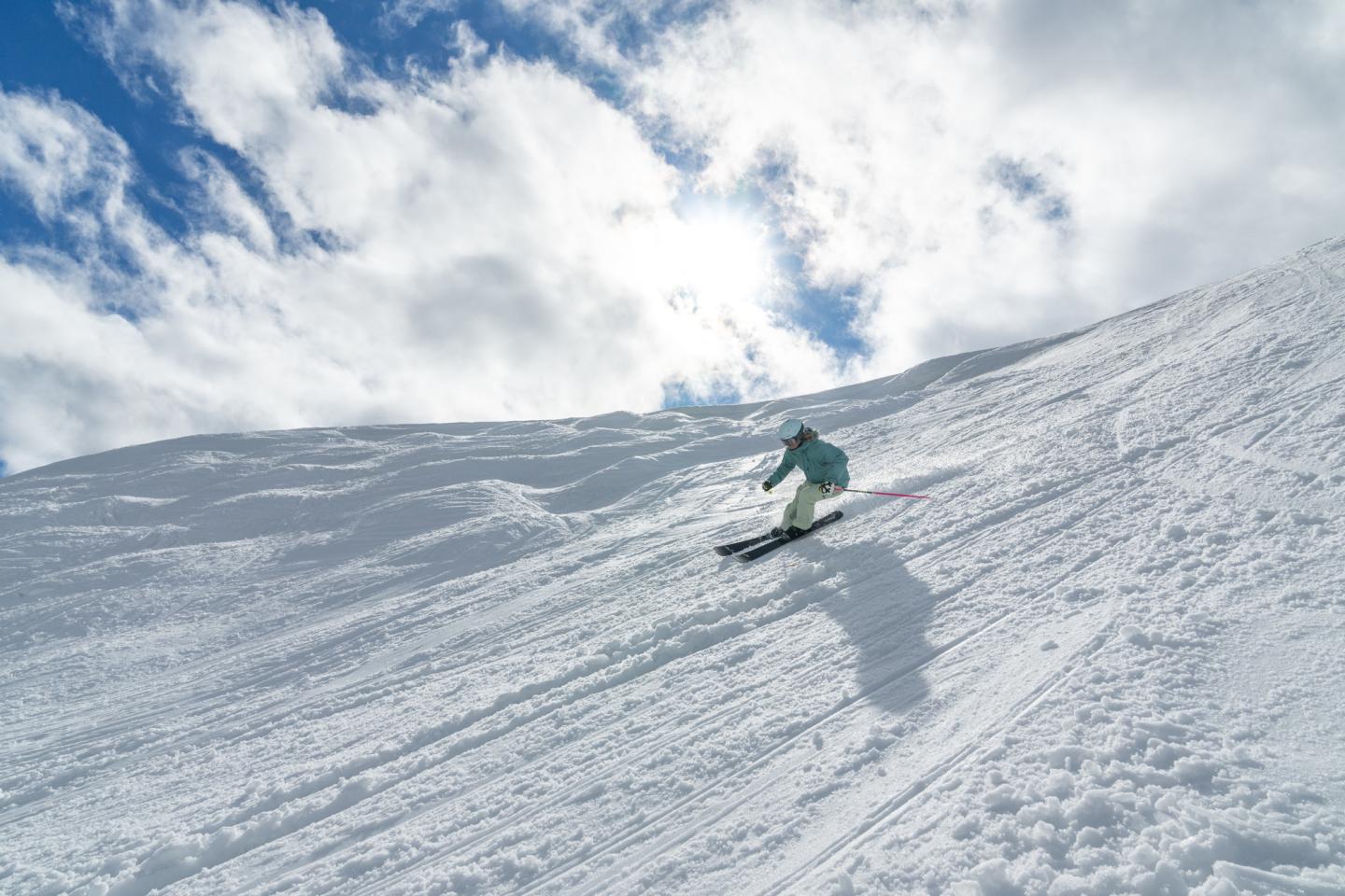 Skier descending snowy slope under blue sky and clouds.