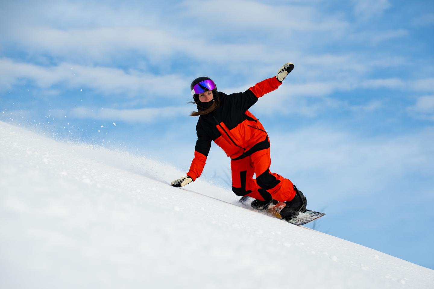 Snowboarder in red and black gear glides down a snowy slope under a blue sky.