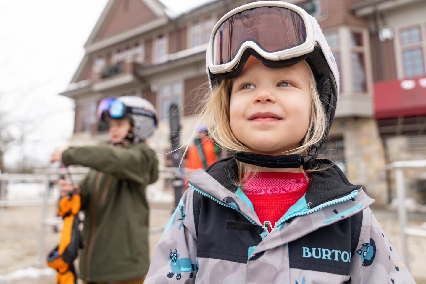 Child smiling in ski gear, standing outside a lodge.