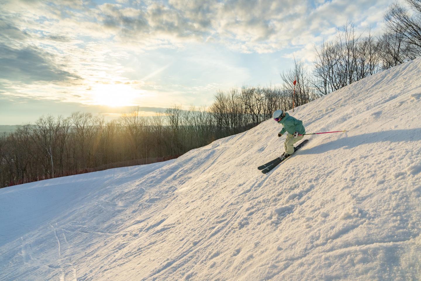 Skier on snowy slope at sunrise, trees in background.