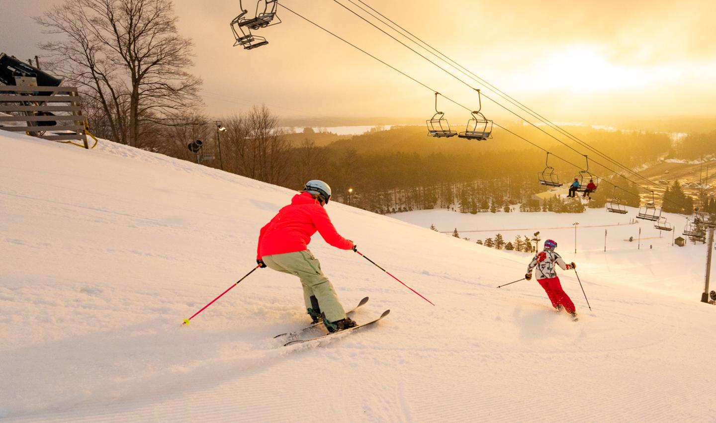 Skiers descending a snowy slope at sunset with a chairlift above.
