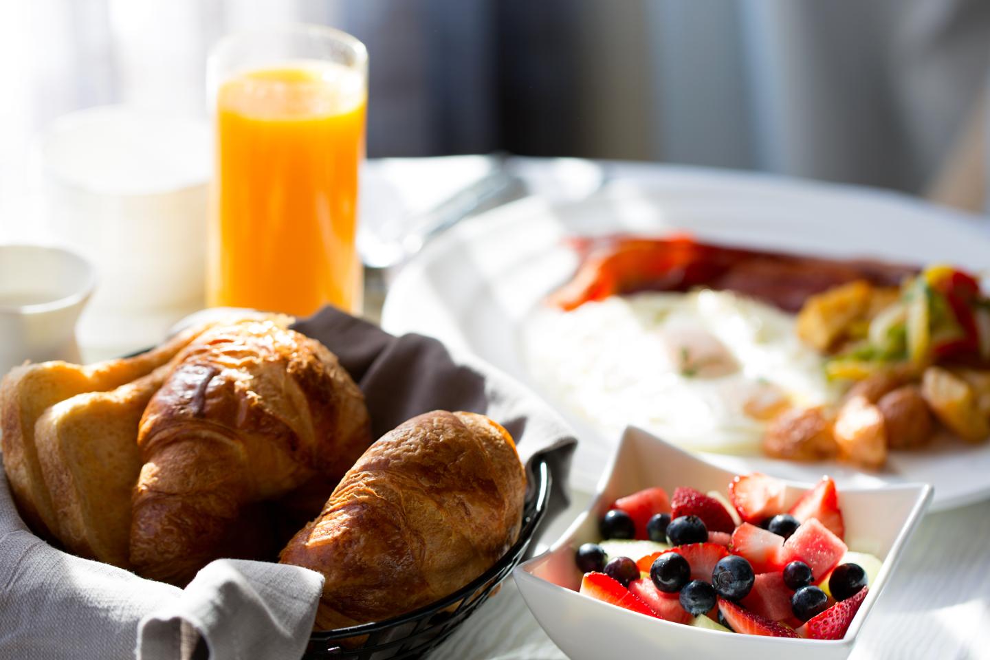 Croissants, fruit bowl, eggs, bacon, and orange juice on a table.