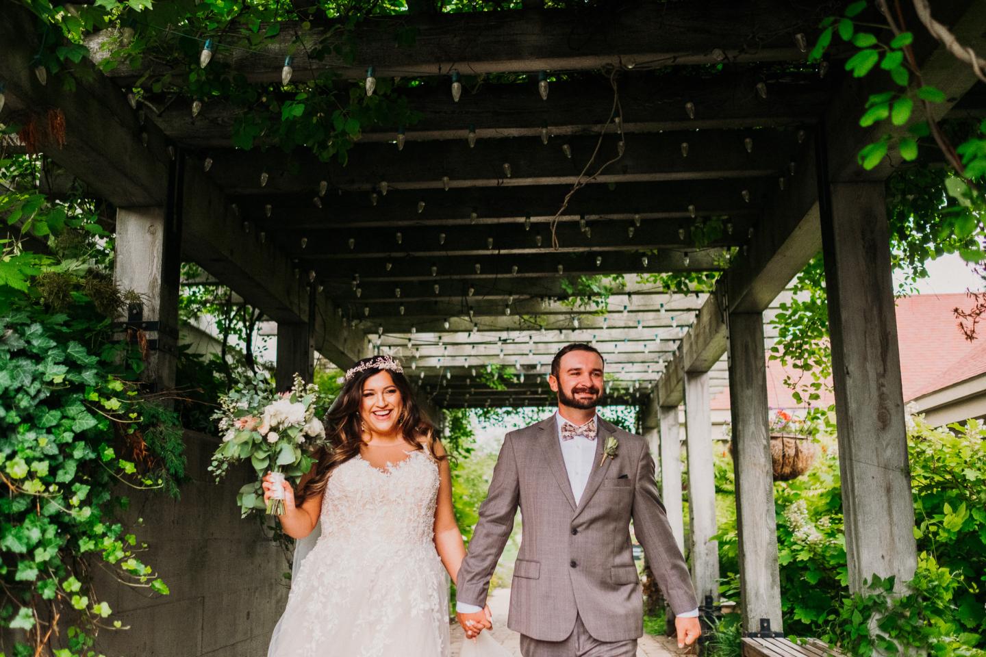 Bride and groom holding hands under a vine-covered pergola, smiling.