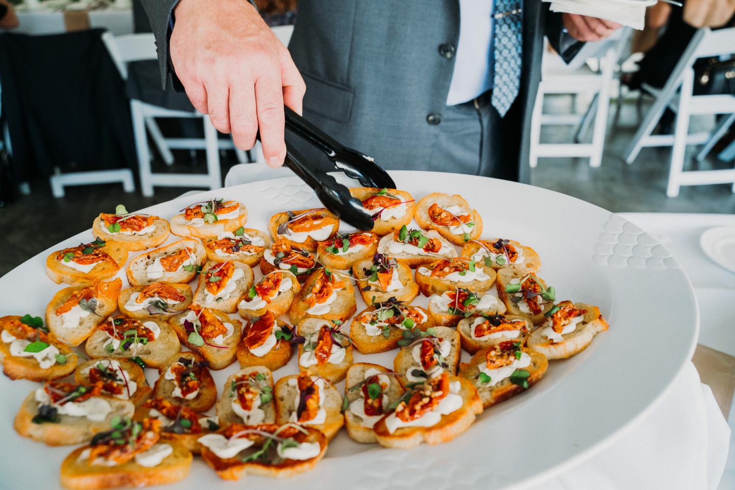 Person picking bruschetta from a platter with tongs at an event.