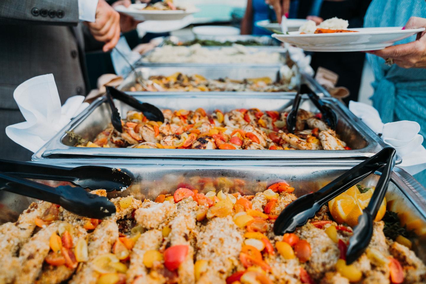 Buffet with colorful dishes and tongs, people serving food onto plates.