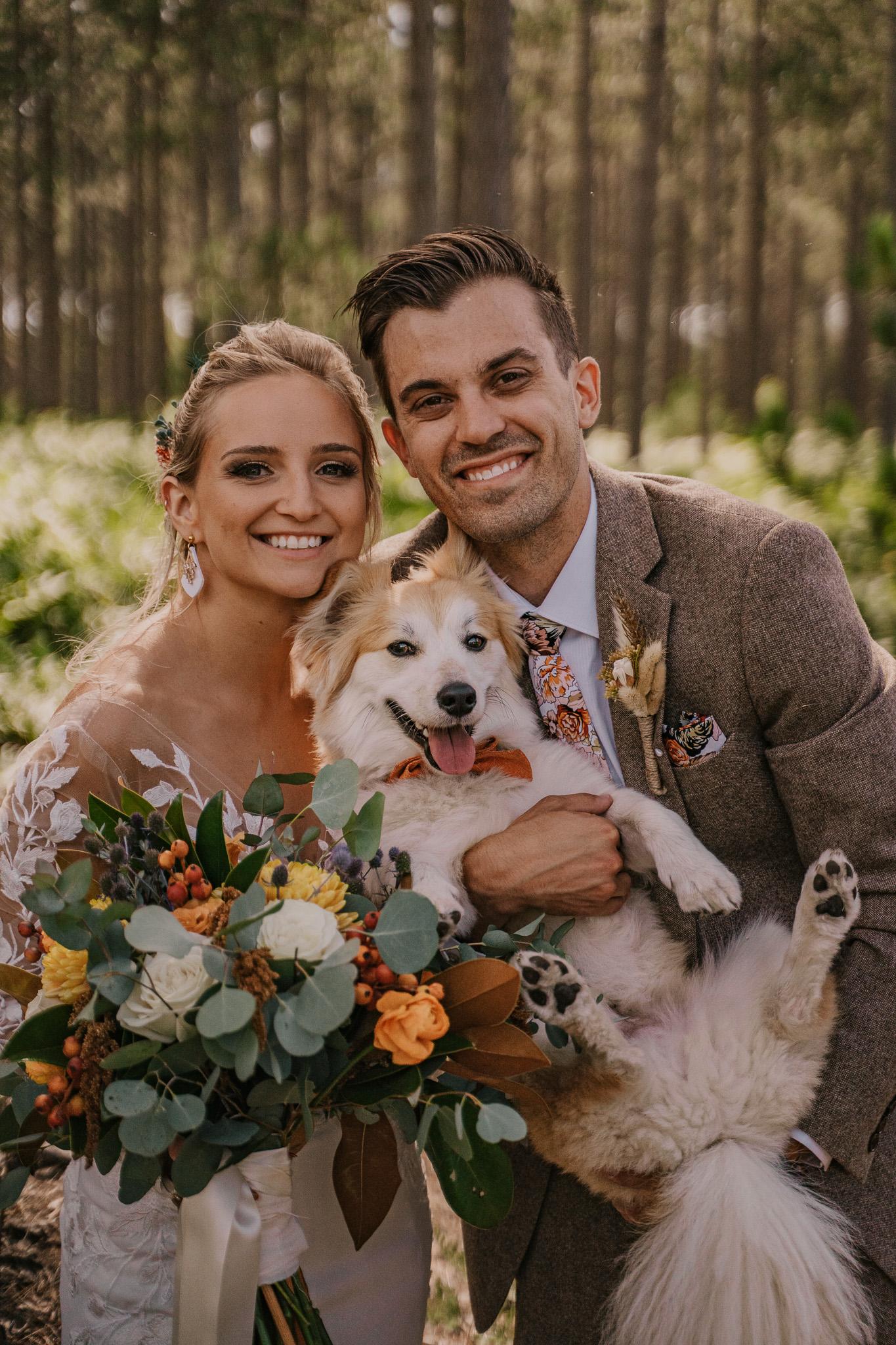 Bride and groom smiling, holding a happy dog, surrounded by trees.