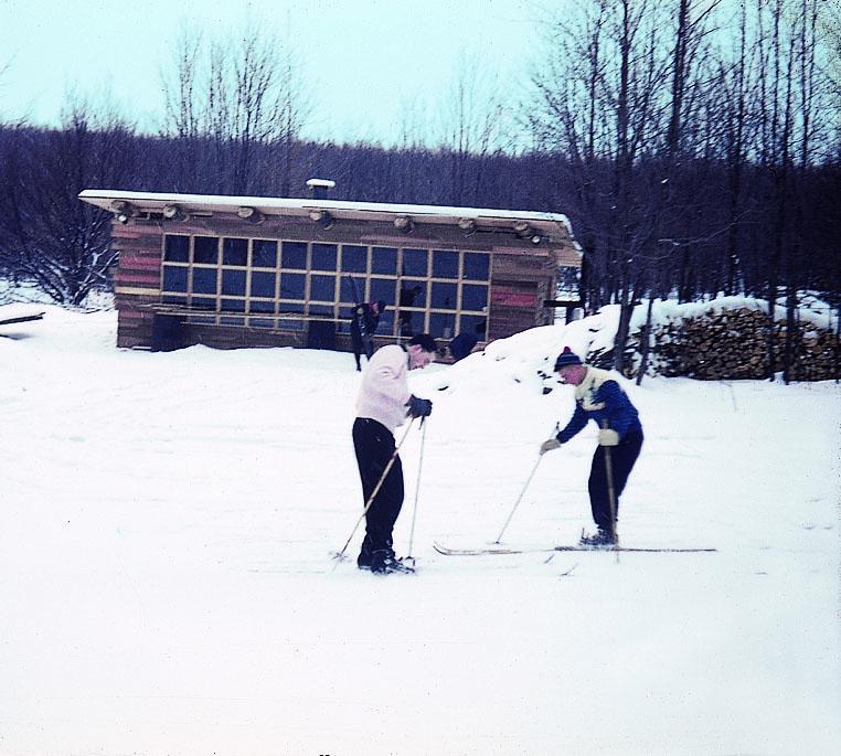 Two people cross-country skiing near a wooden cabin in snowy landscape.