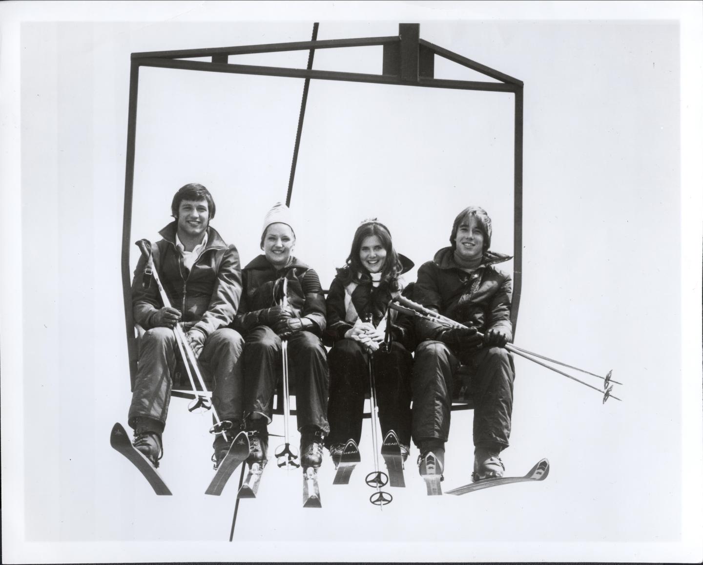 Four people on a ski lift, holding ski poles, smiling. Black and white photo.
