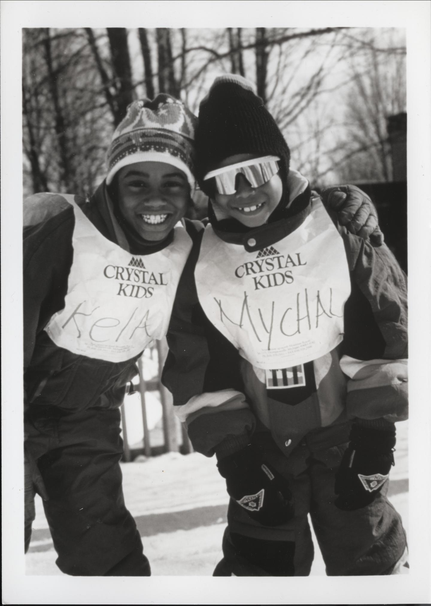 Two kids smiling in winter gear and Crystal Kids vests outdoors.