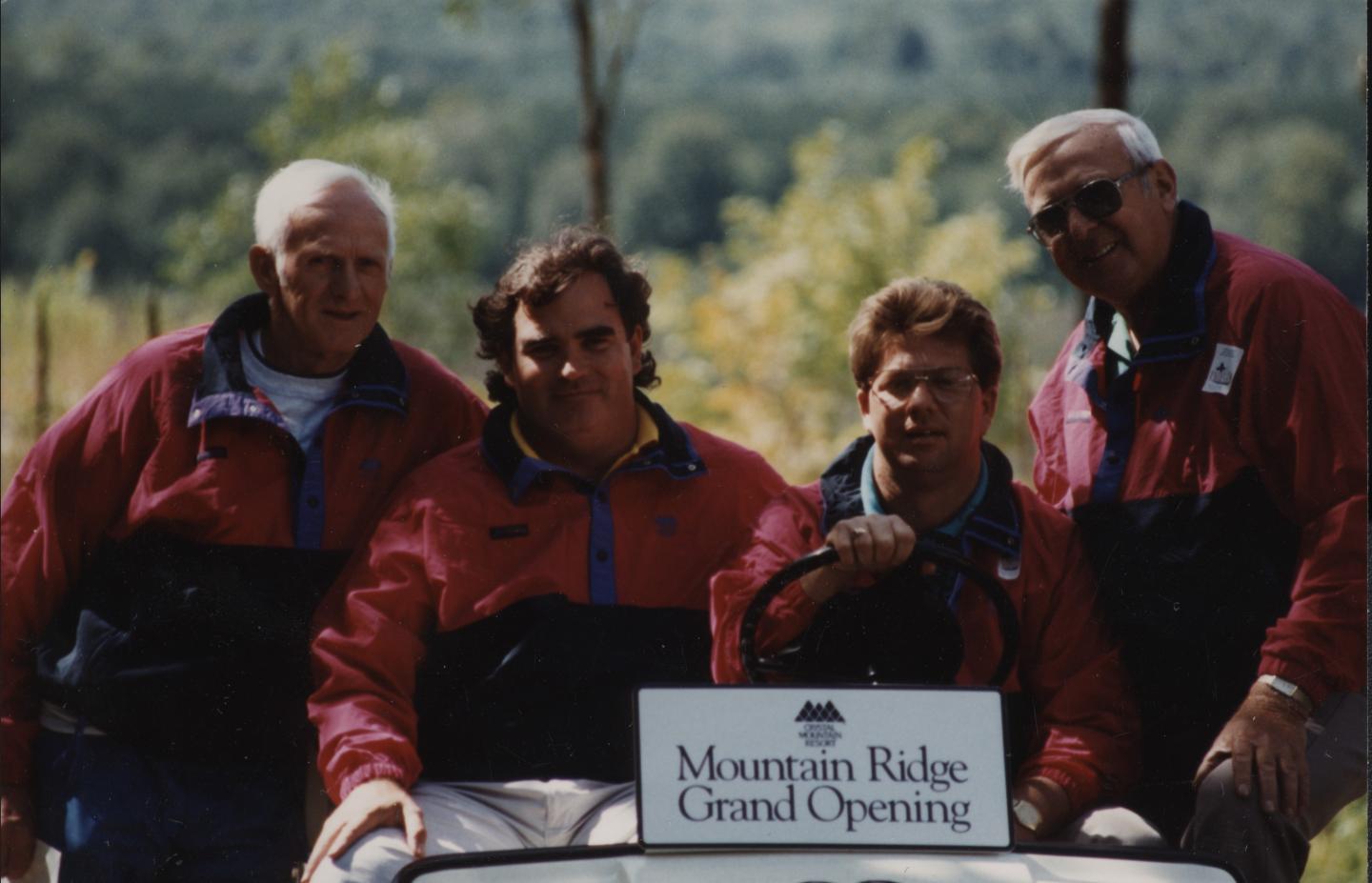 Four men in matching jackets at outdoor event with mountain backdrop.