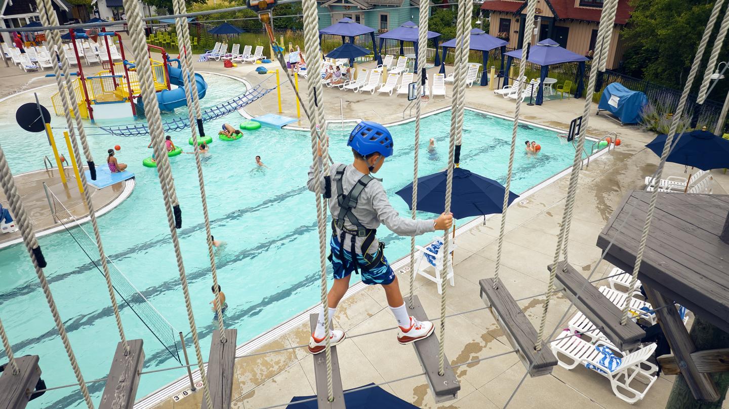 Child in helmet on ropes course near a pool.