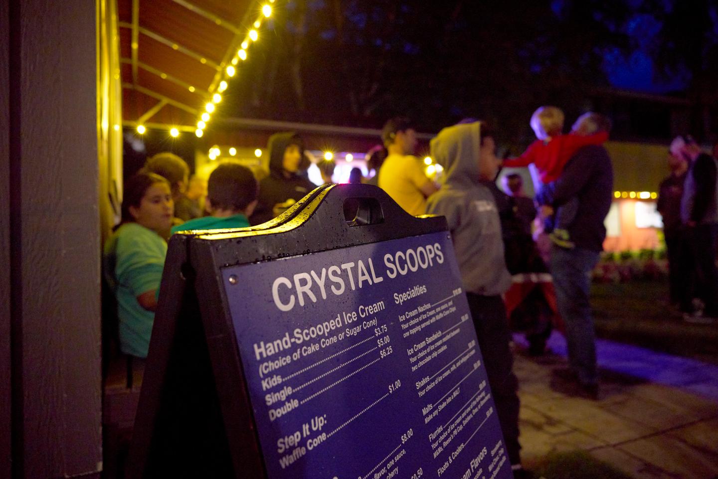 Nighttime ice cream shop with menu board and people gathered under string lights.