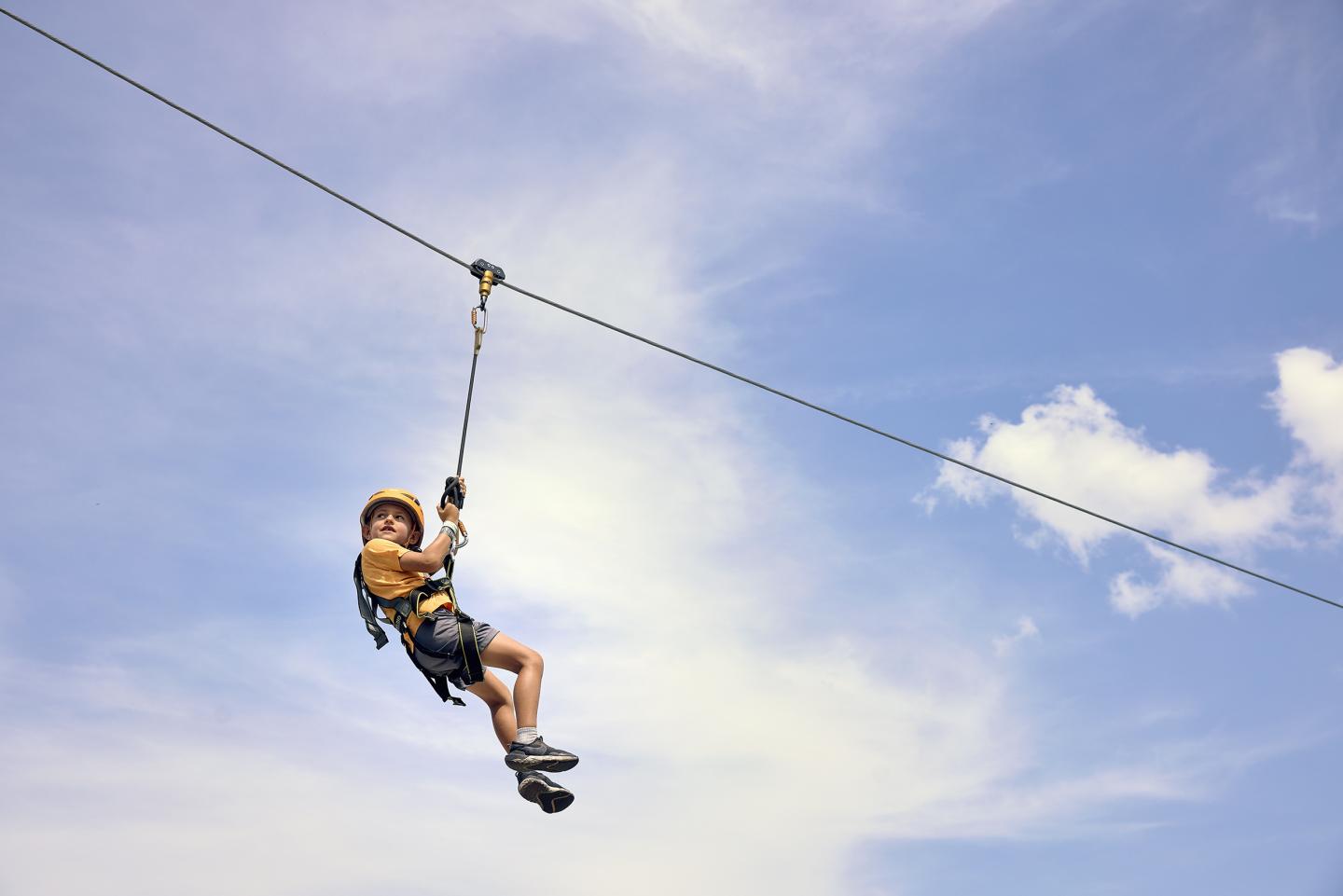 Child ziplining under a blue sky with clouds.