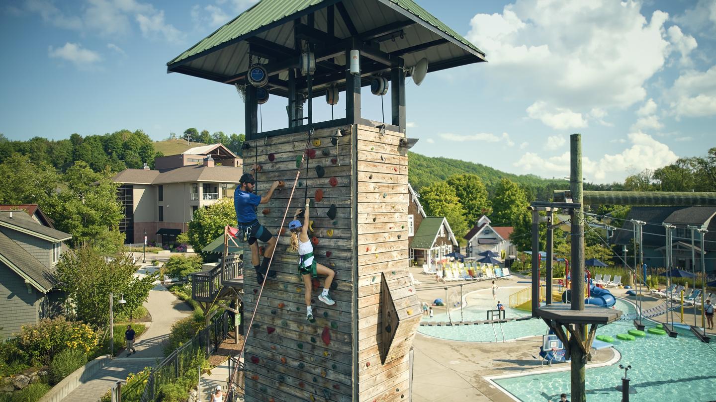 Rock climbing tower beside a swimming pool, surrounded by buildings and trees under a blue sky.