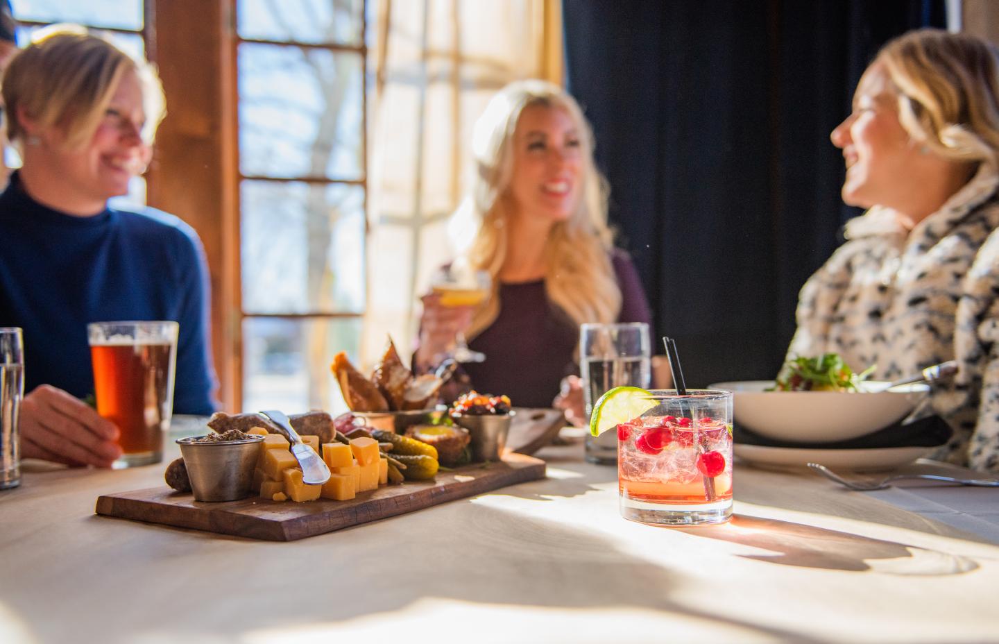 Three people enjoying drinks and a meal at a sunlit restaurant table.