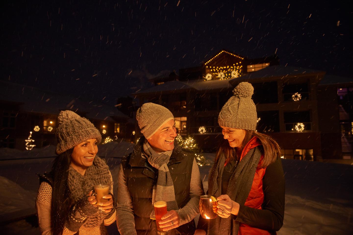 Three people in winter hats enjoying hot drinks outside in a snowy evening setting.