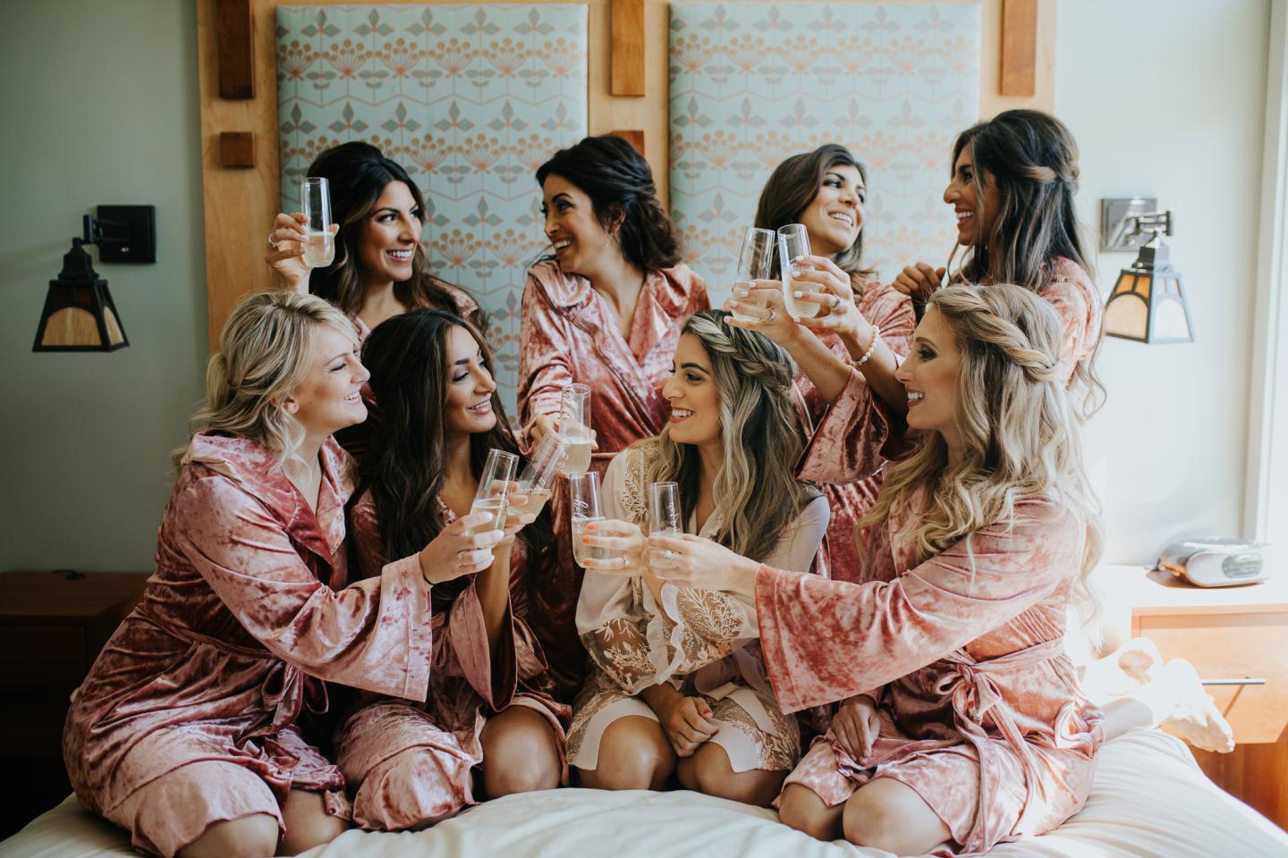 Women in pink robes smiling and toasting with champagne on a bed.