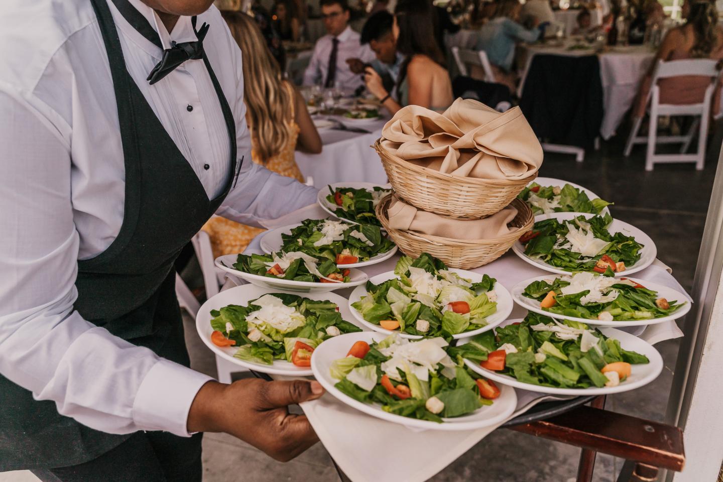 Waiter carries a tray of salads and bread baskets at a formal event.