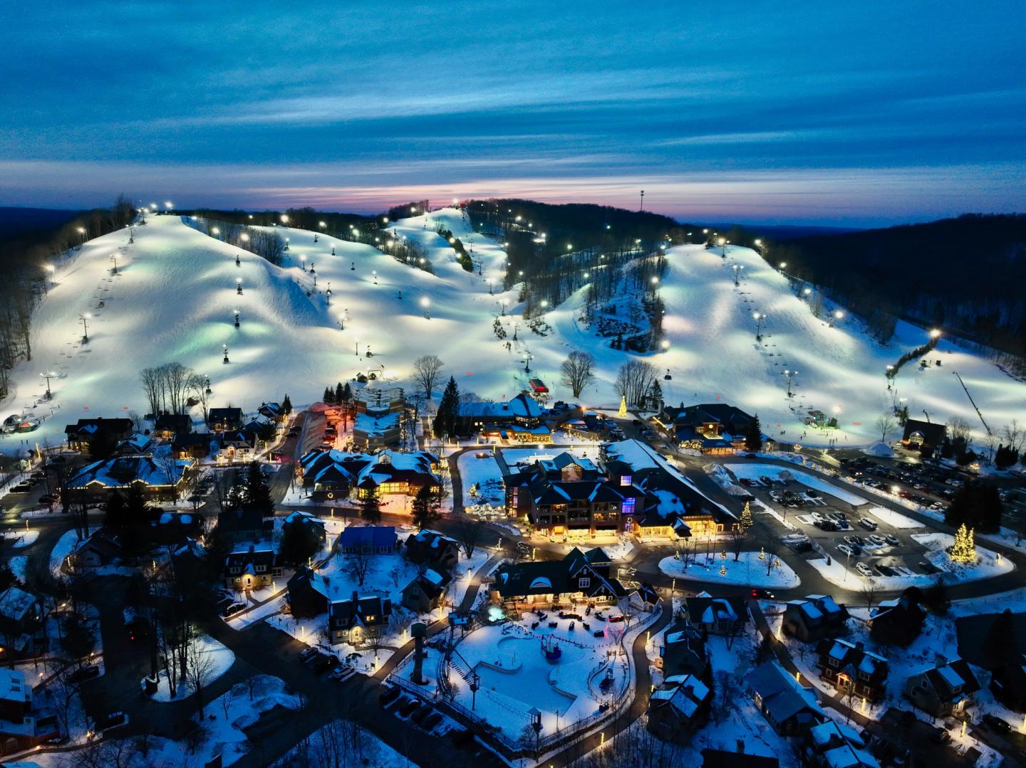 Snowy ski resort lit up at twilight with hills and buildings.