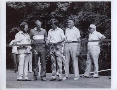 A group of five men standing on a road in a forest setting, dressed in casual attire.