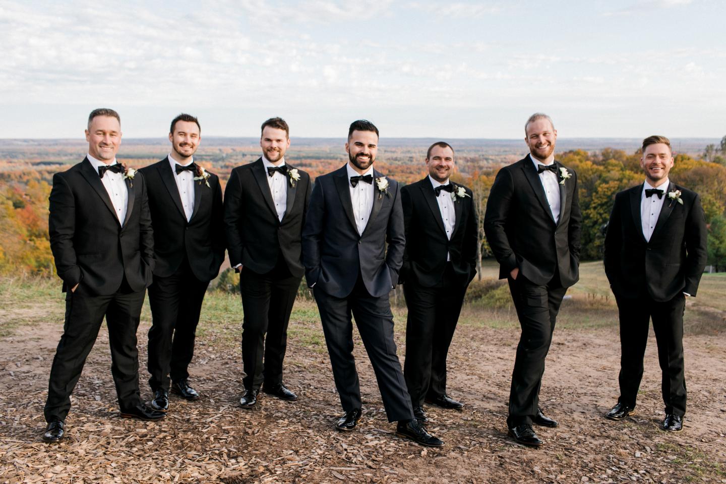 Seven men in tuxedos smiling outdoors on a clear day.