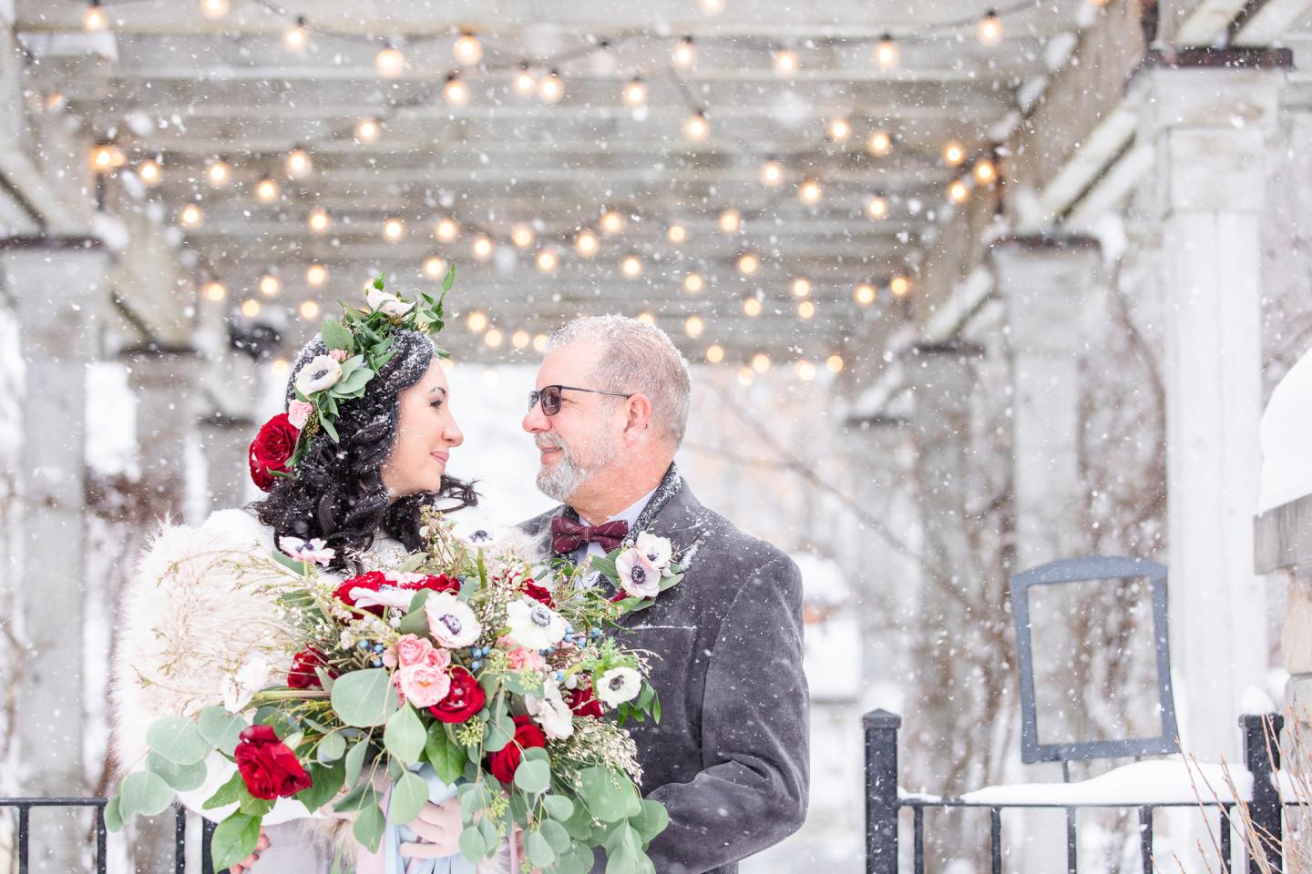 Couple smiling with winter floral bouquet under string lights in snowy setting.