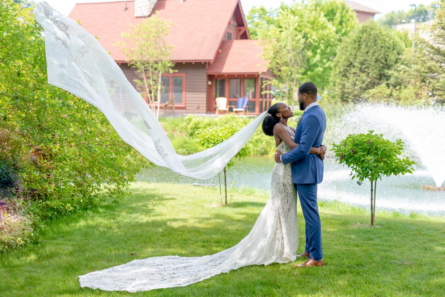 Wedding couple kissing by a pond, veil flowing, greenery and fountain in background.