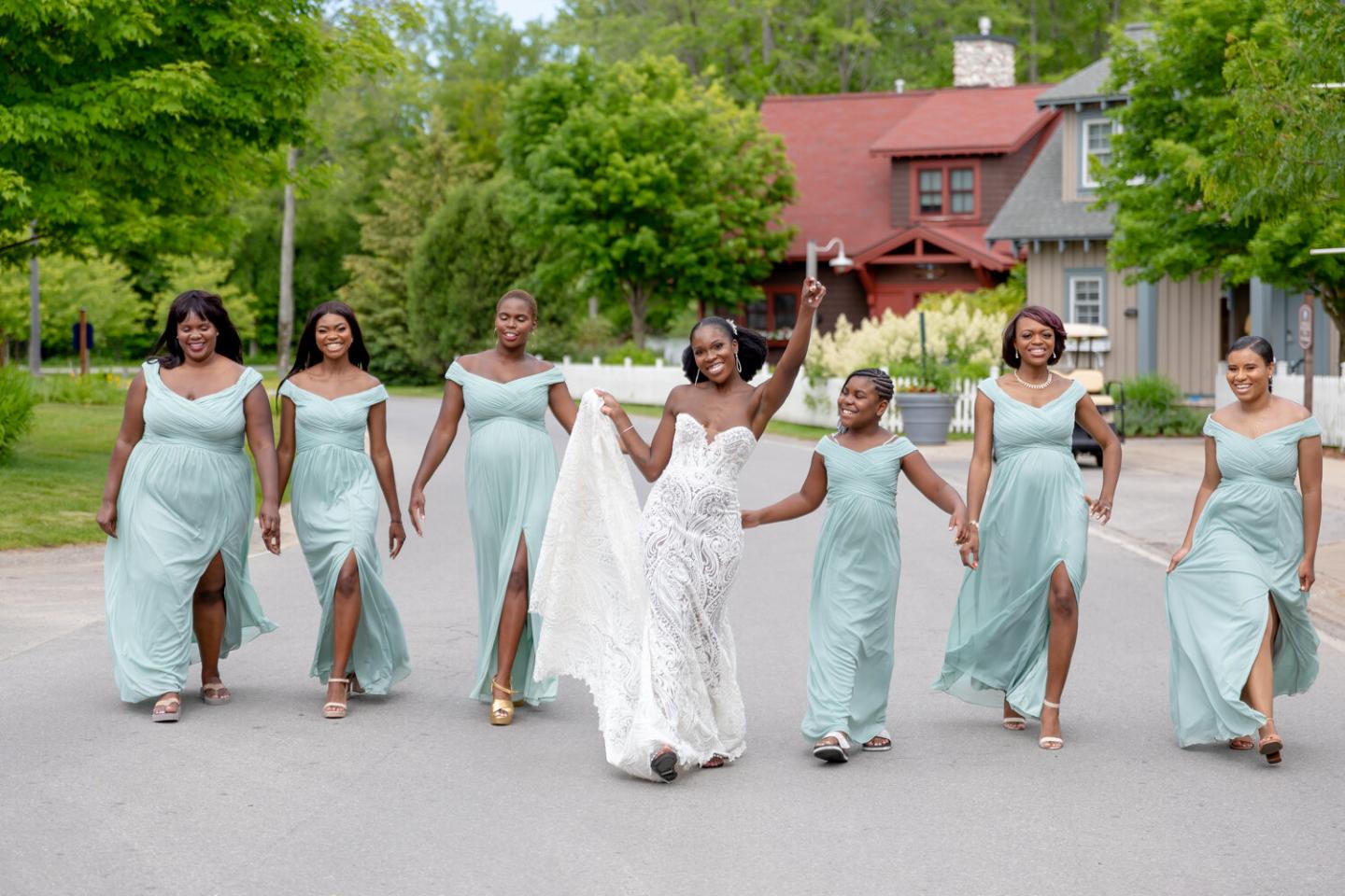Bride in white gown joyfully walks with bridesmaids in mint dresses on a street lined with trees.