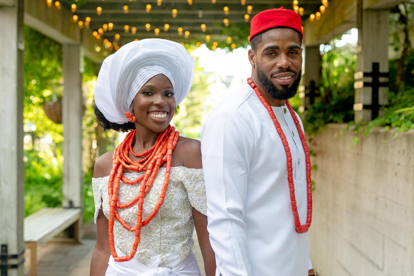 Couple in white attire and red accessories under string lights, smiling warmly.
