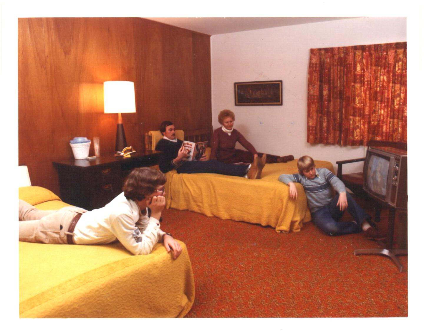 Four people relax in a retro-style hotel room with yellow bedspreads and an old TV.