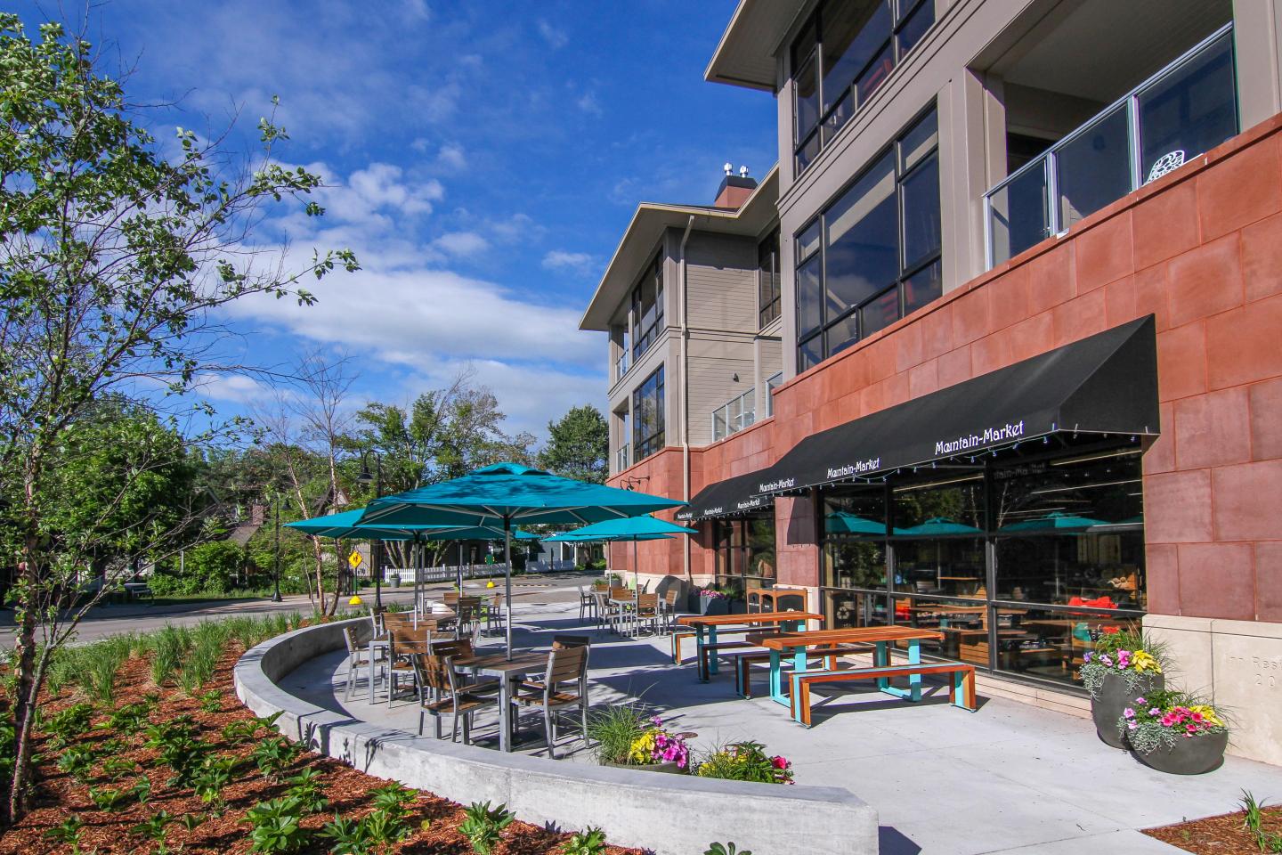 Outdoor cafe patio with tables, umbrellas, and a landscaped garden on a sunny day.