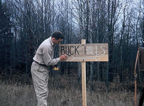 Man adjusting a "Buck Hills" sign in a forest clearing.