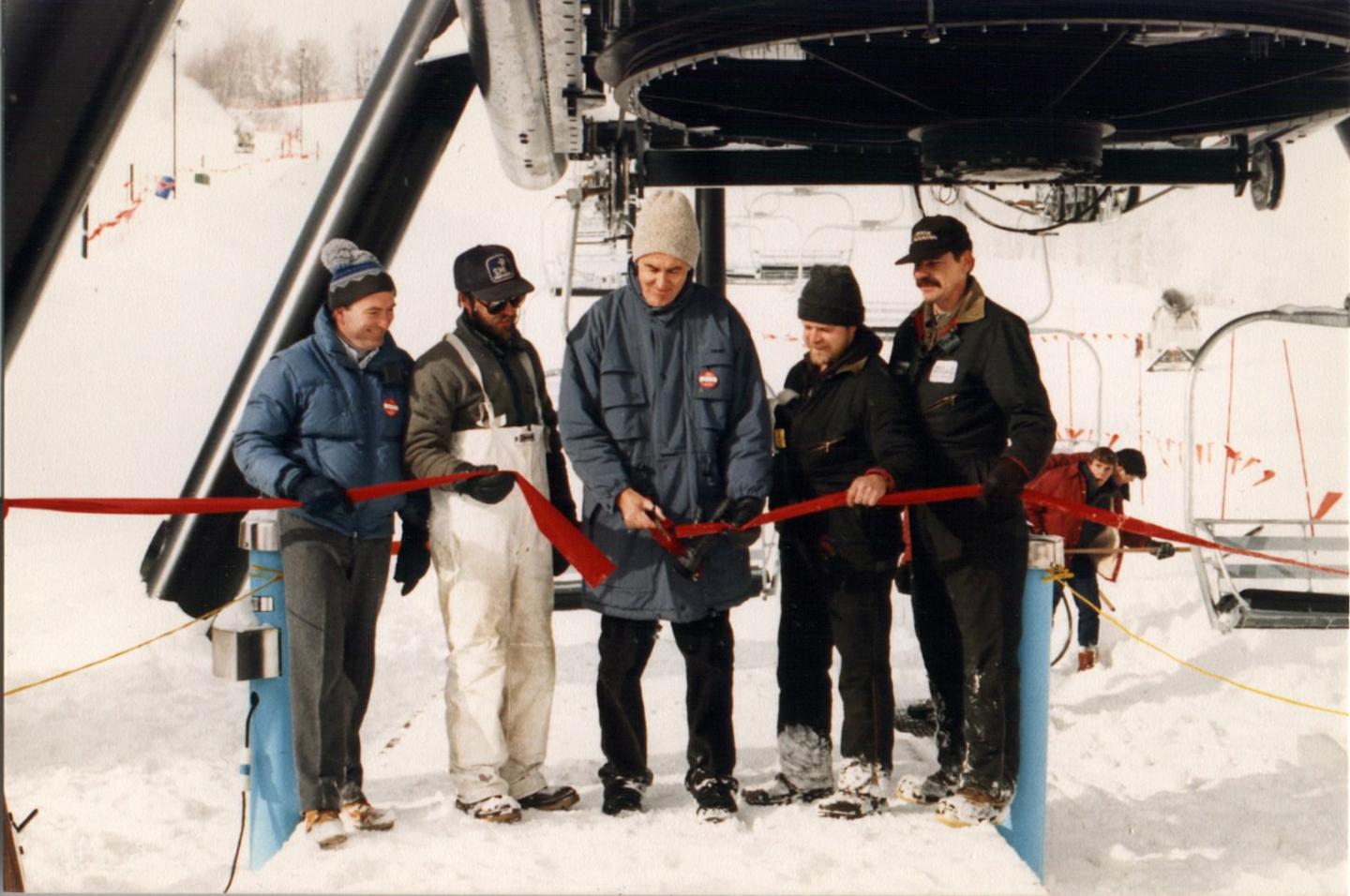 Man cutting red ribbon at snowy ski lift ceremony, five people present.