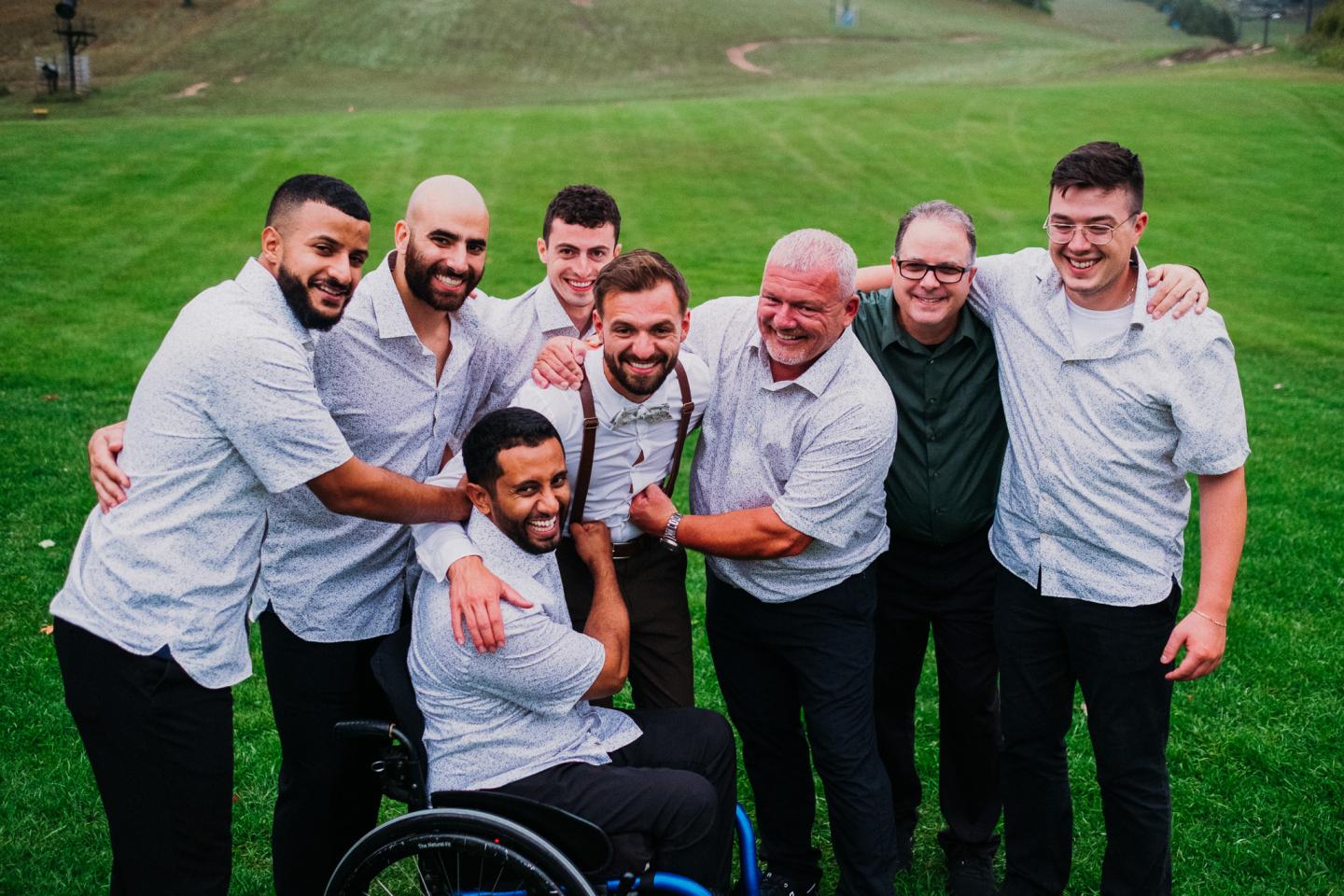 A group of eight smiling men pose on a grassy field, one in a wheelchair.