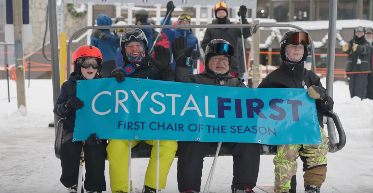 Four people in snow gear on a ski lift, smiling and holding a banner.