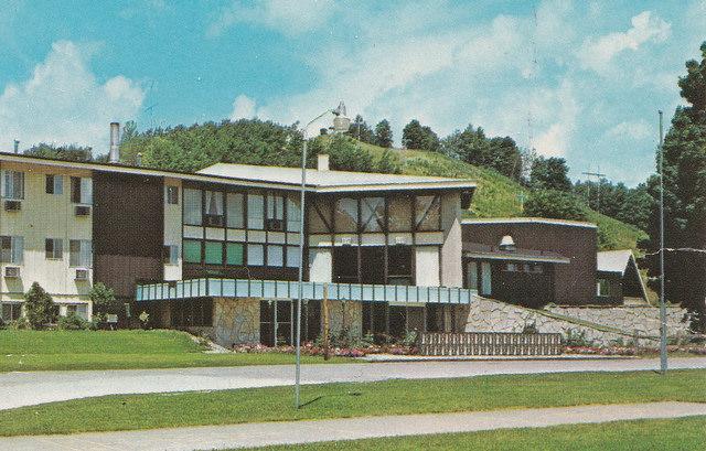 The Lodge with glass facade, surrounded by green hills and trees, under blue sky.
