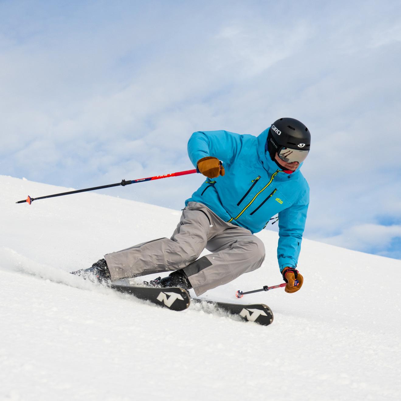 Skier in blue jacket descending a snowy slope under a cloudy sky.