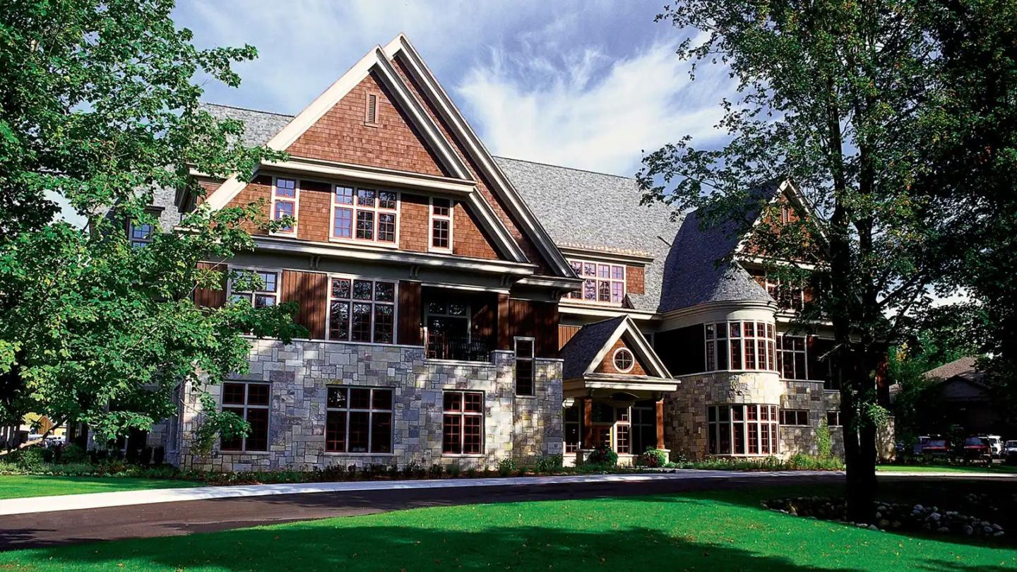Large brick and stone house surrounded by trees and lawn under a blue sky.