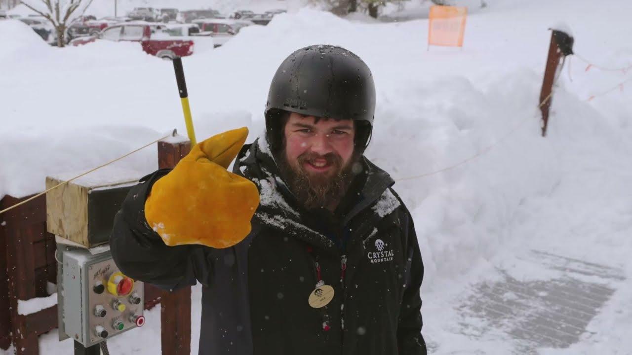 Man in helmet and yellow gloves giving thumbs up in snowy setting.