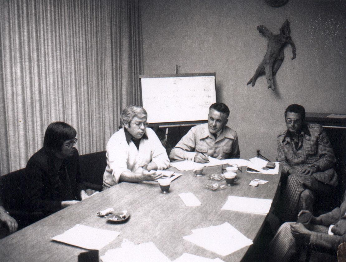 Five people seated around a table in a meeting room with papers and a whiteboard.