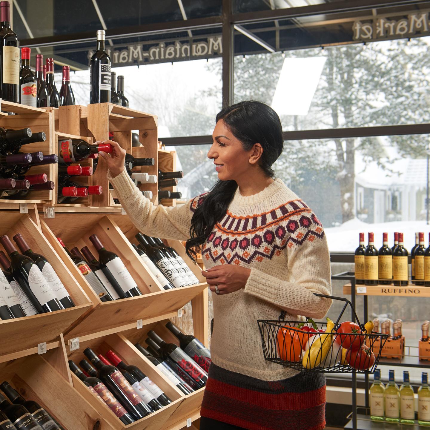 Shopper in a sweater selecting wine from a wooden shelf in a market.