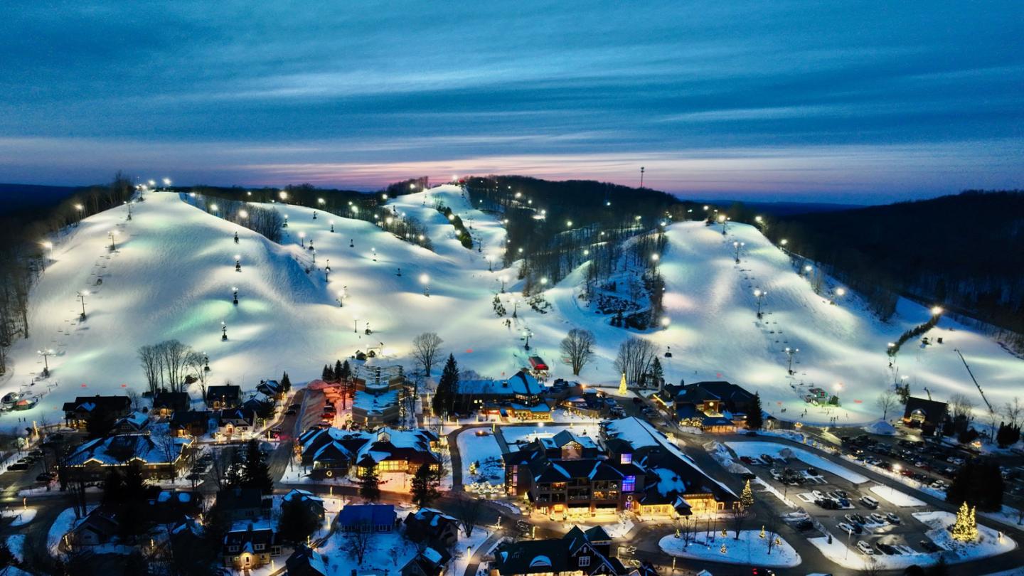 Snowy ski resort at dusk, slopes lit with lights, small village below.